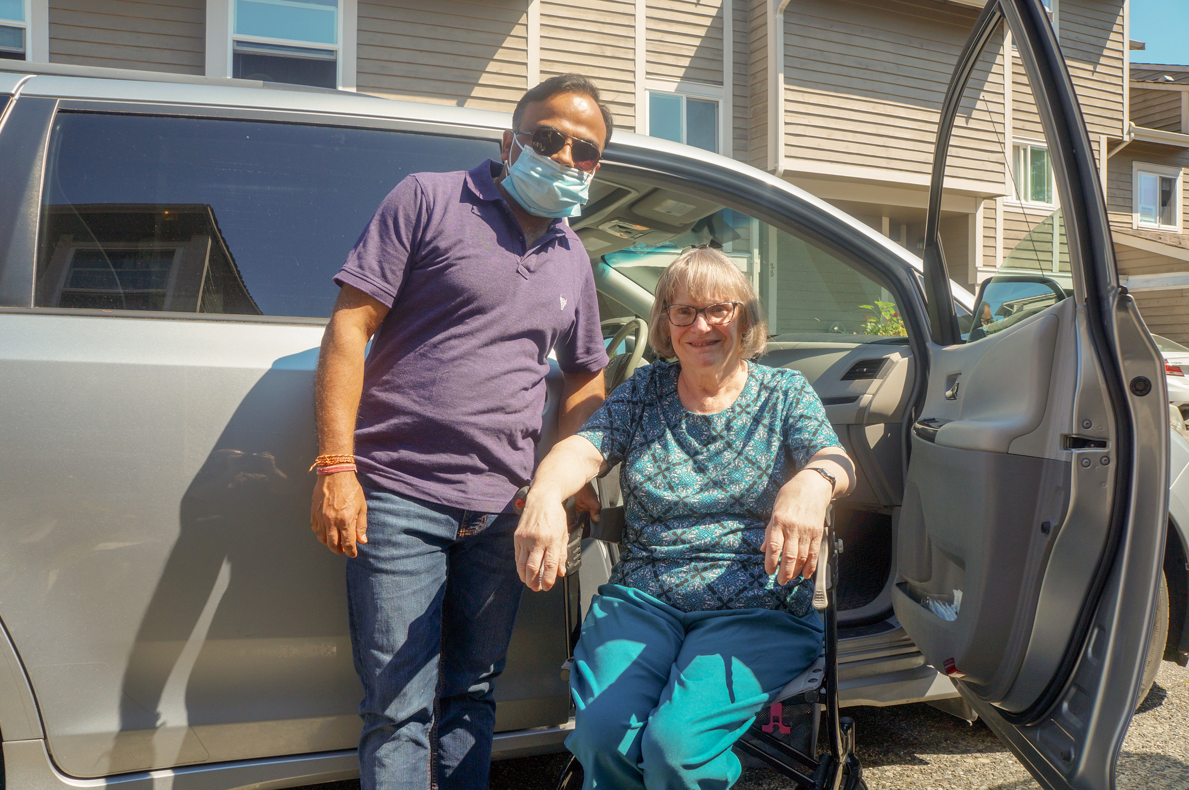 A man in a facemask standing next to an older lady, who's sitting in a walker