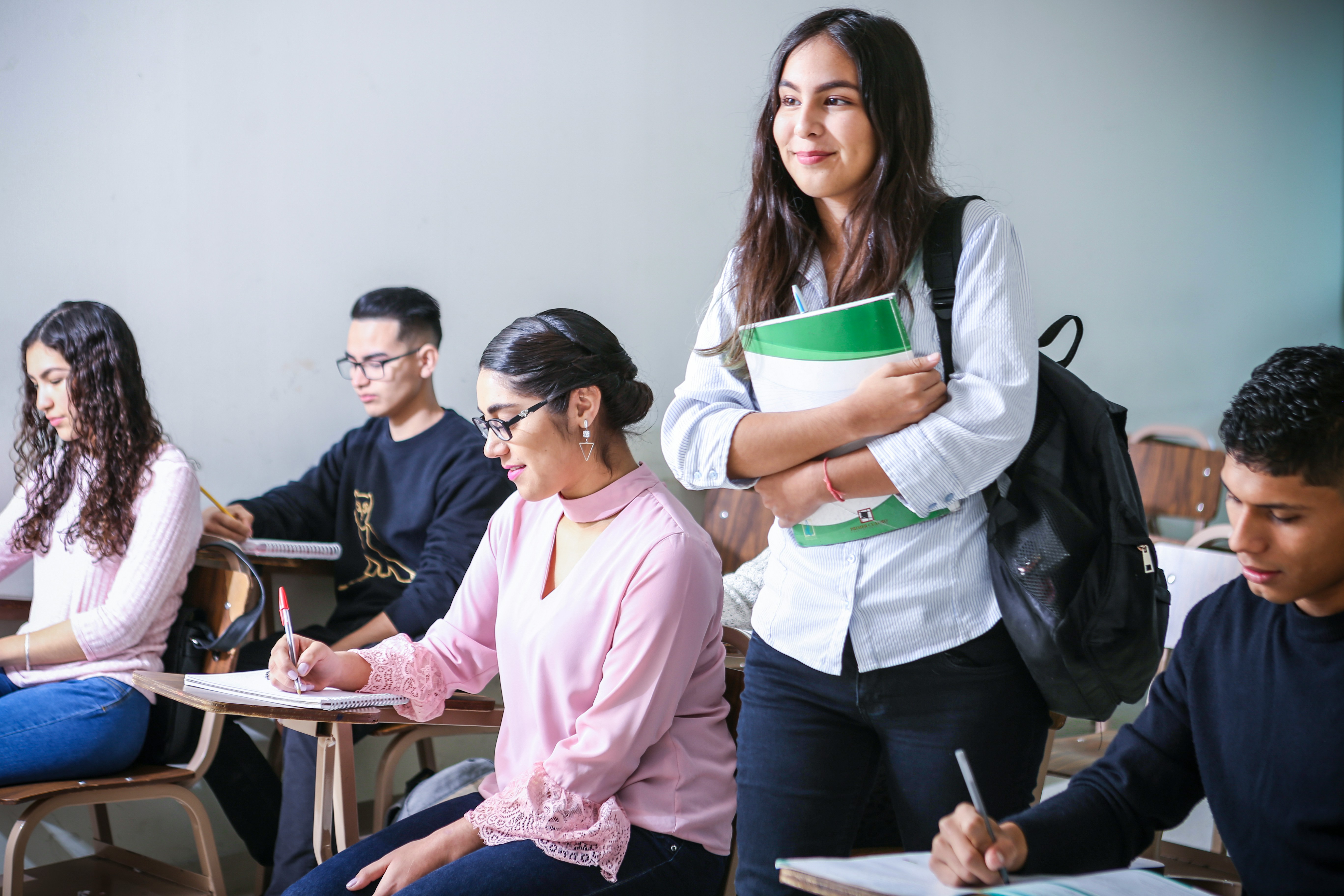 A female student holding books and a backpack, while walking amongst fellow students seated at desks