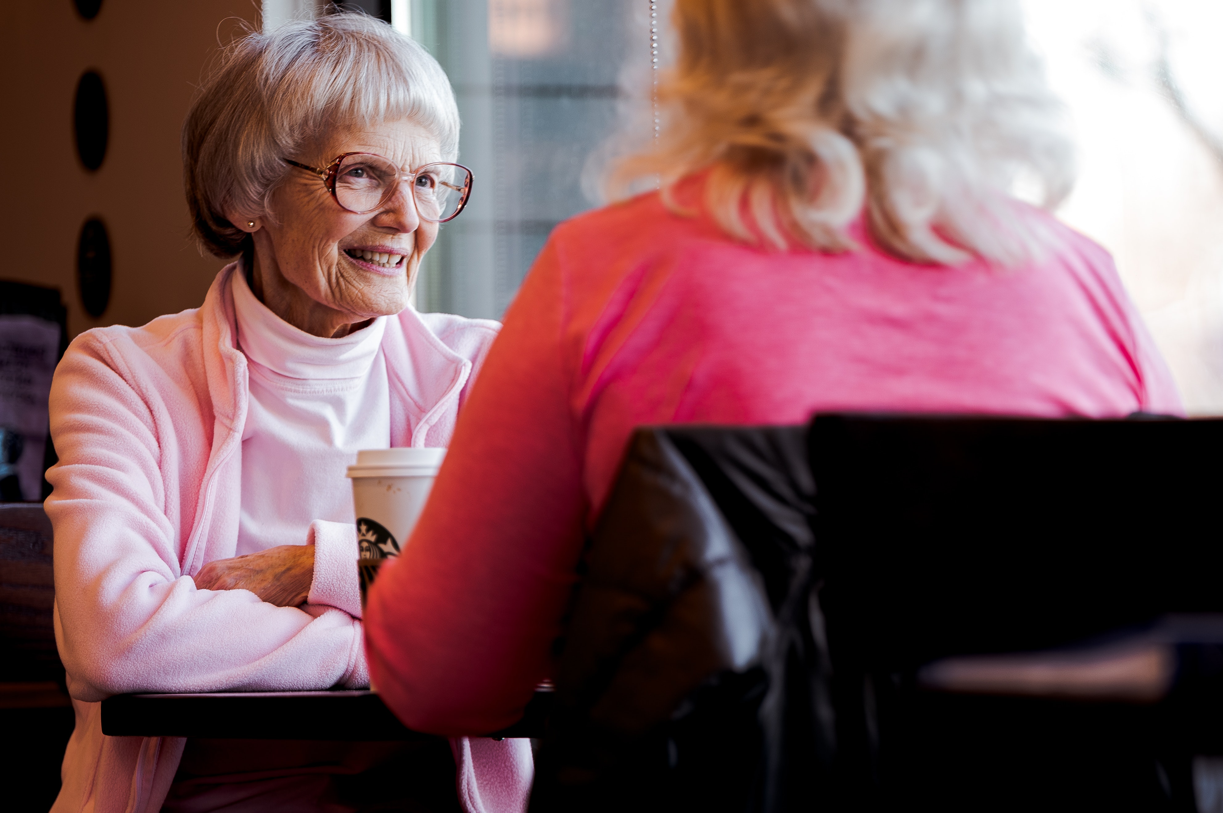 Two older ladies in pink shirts sitting in a café, next to a window